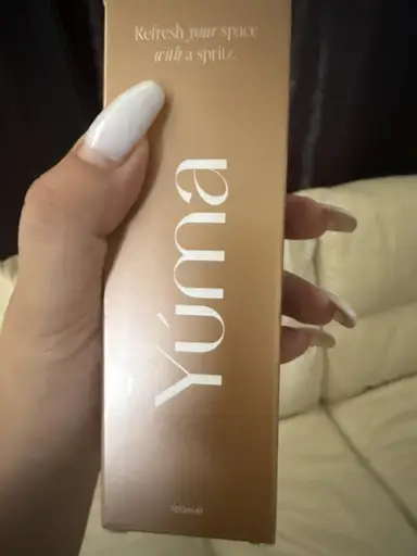 A hand with white nails holding the bronze YUMA packaging in front of a sofa.