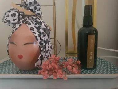 A dark YUMA room spray bottle next to a decorative head wearing a leopard-print turban, surrounded by pink flowers.