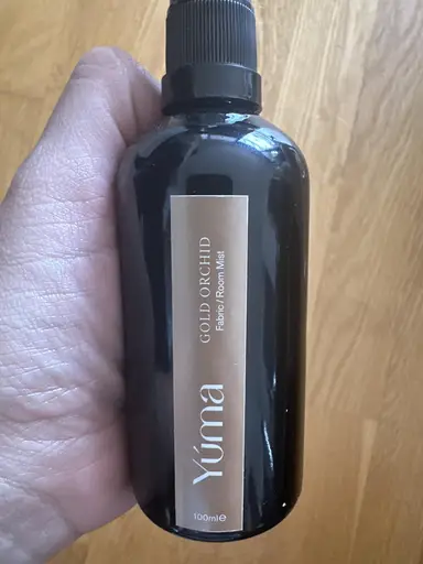 A hand holding the YUMA Gold Orchid Room Mist black bottle in front of a wooden floor.