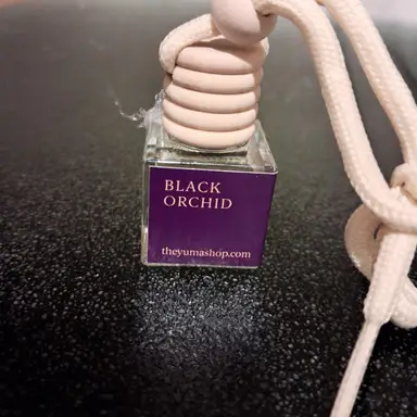Small glass Black Orchid car diffuser with a wooden cap and beige cord on a black surface.