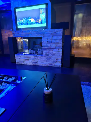 A reed diffuser on a dark table with a stone fireplace and TV in the background, lit by blue lighting.