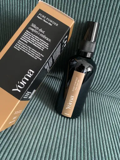 A black bottle of YUMA Pure Powder Room Mist next to its packaging, on a green striped fabric.