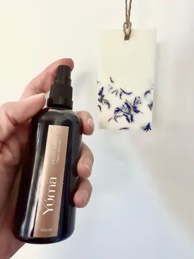 A hand holding YUMA Fresh LINEN room spray next to a hanging scented candle.