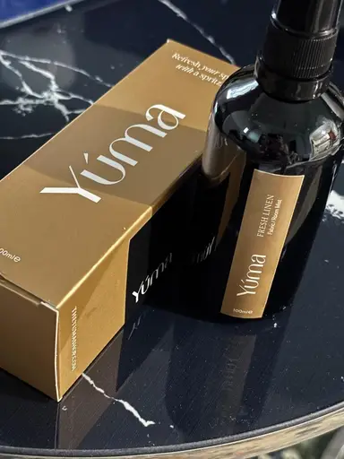 A dark bottle of YUMA Fresh Linen room spray beside its gold packaging, placed on marble.