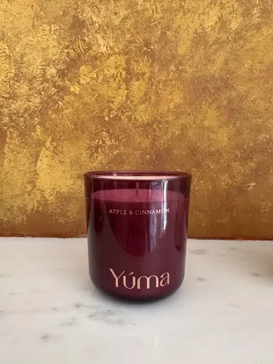 YUMA Apple & Cinnamon scented candle in a burgundy glass jar in front of a gold wall.