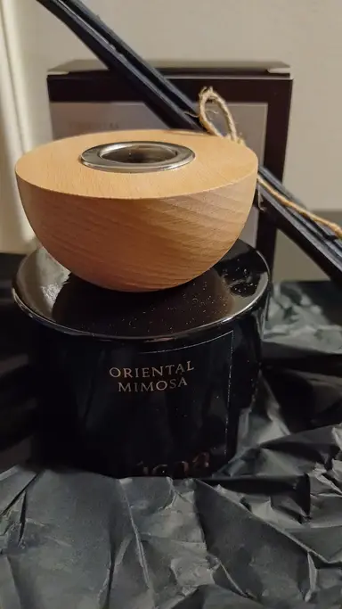 Black glass Oriental Mimosa reed diffuser with a wooden cap and black reeds, set in crinkled paper.