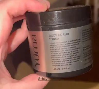 A hand holding the YUMA TONKA Body Scrub in a black jar with a silver label.