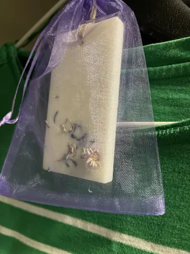 A white scented wardrobe sachet candle with dried flowers, inside a purple organza pouch on green fabric.