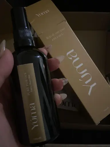 A hand holding YUMA Pure Powder room mist next to its beige packaging.