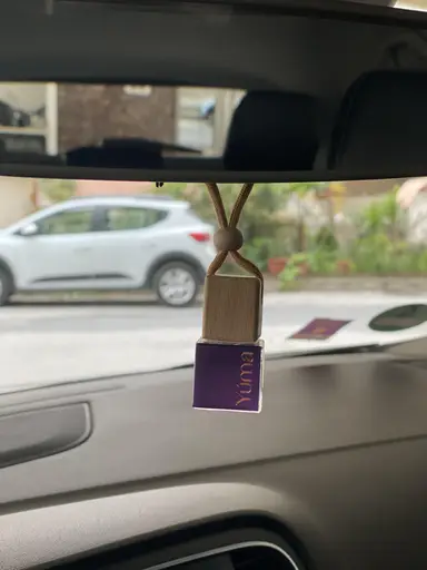 A small YUMA glass car fragrance diffuser with a purple label and wooden stopper, hanging from a rear-view mirror.