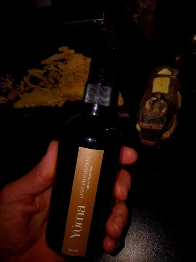 A hand holding the YUMA Luxe Philoxenia Room Mist in a dark bottle with a gold label.