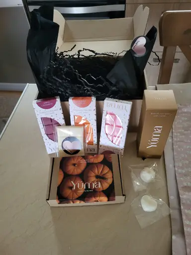 Open YUMA gift box containing wax melts, a room fragrance spray, and black decorative tissue paper.