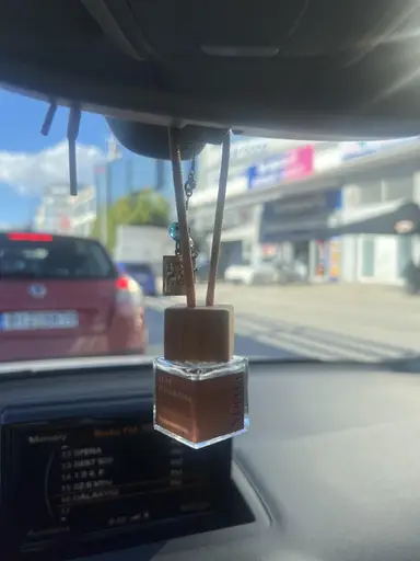 YUMA Luxe Philoxenia car diffuser hanging from a rearview mirror, with a road and cars visible in the background.