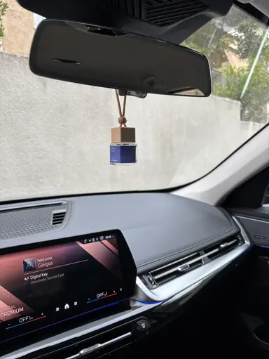 A YUMA Fresh Linen hanging car freshener with a wooden stopper on a vehicle's interior mirror.