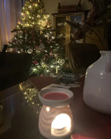 A white ceramic wax burner with a lit tea light and a red candle, in front of a decorated Christmas tree.