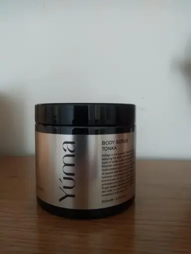 A black YUMA Tonka Body Scrub jar with a silver label, placed on a wooden surface.