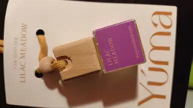 A small glass Lilac Meadow car air freshener with a wooden stopper and yellow cord, resting on white YUMA packaging.