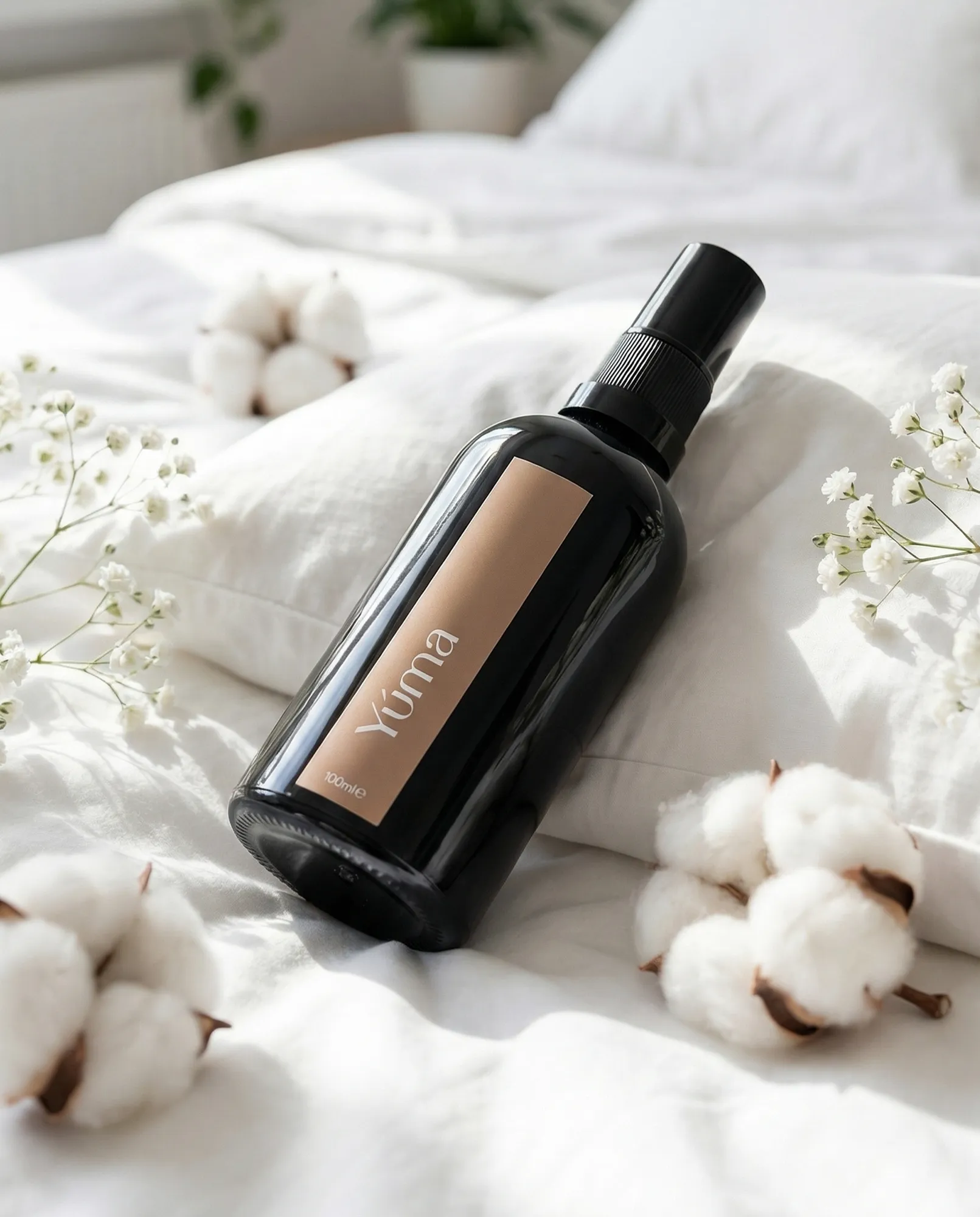 Dark bottle of YUMA Fresh Linen Fabric/Room Mist on white bedding with cotton flowers and gypsophila.