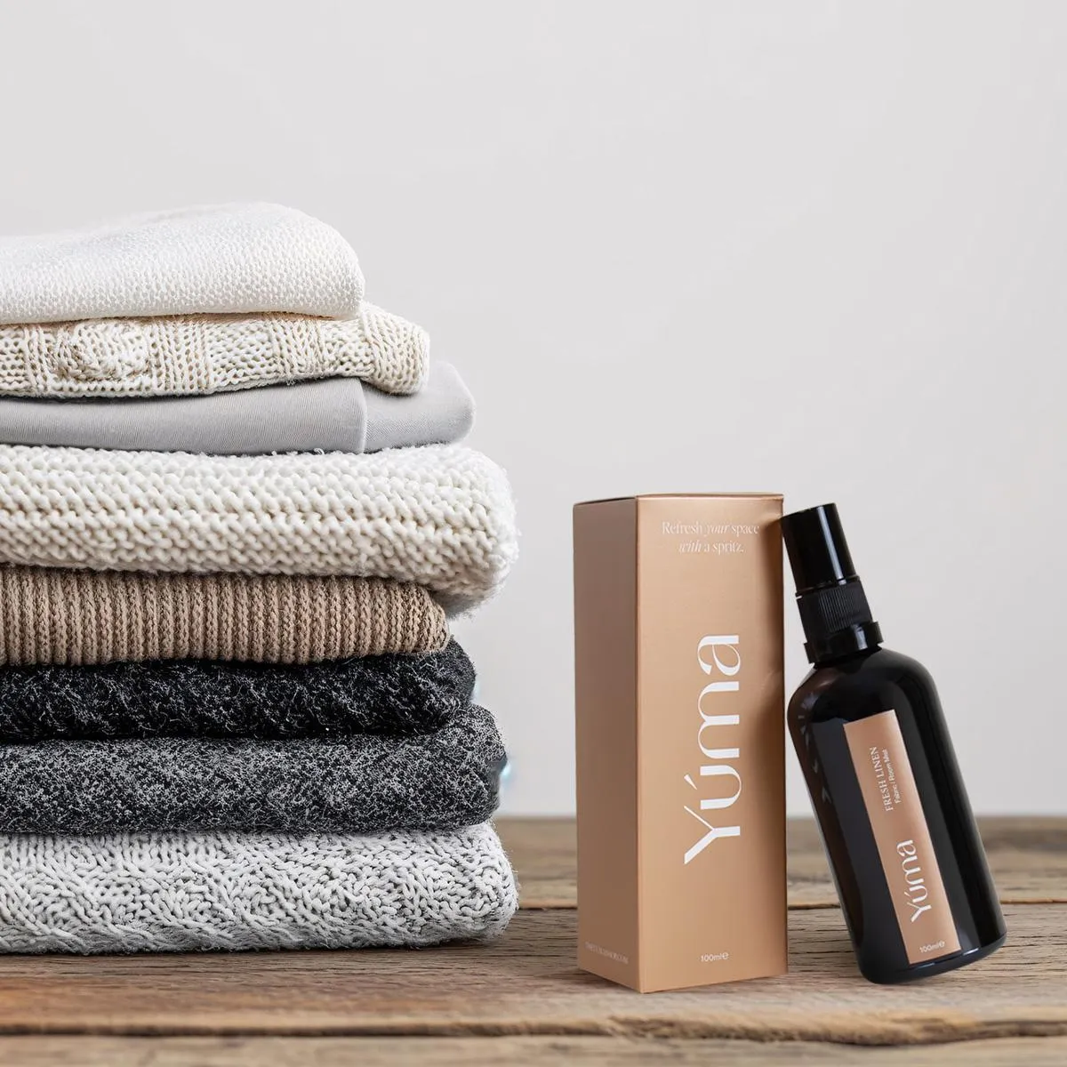 YUMA Amber Tobacco fabric spray next to a stack of folded knit garments on a wooden surface.