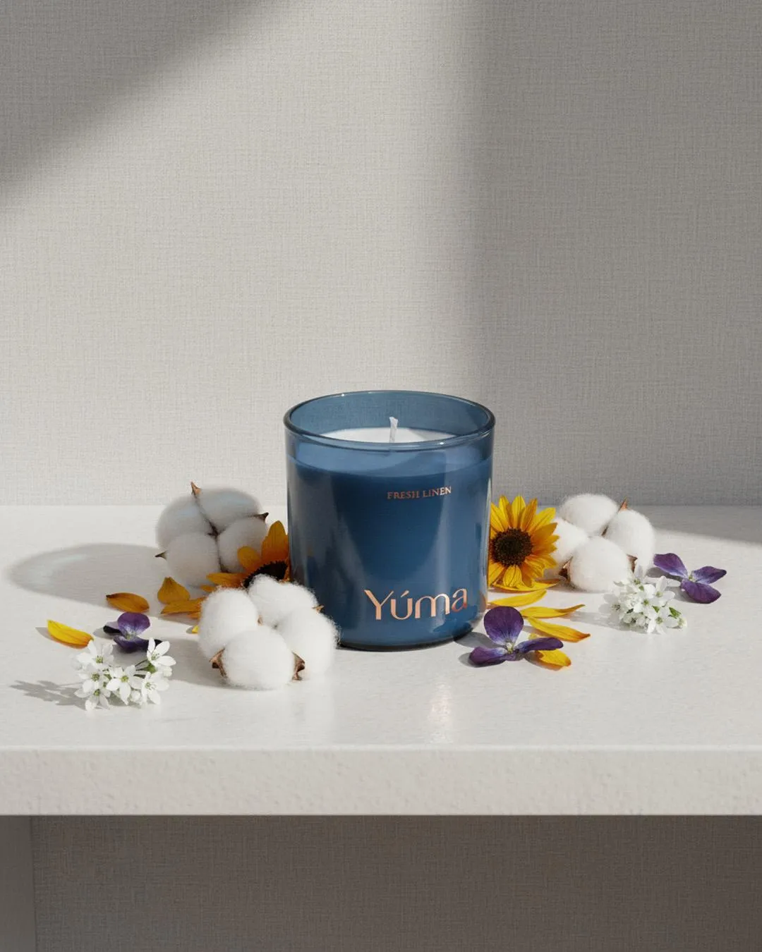 YUMA Fresh Linen candle in a blue glass vessel among cotton flowers and yellow petals.
