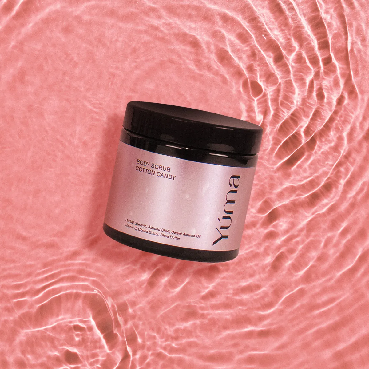 YUMA Cotton Candy Body Scrub in a dark jar resting on pink rippling water.