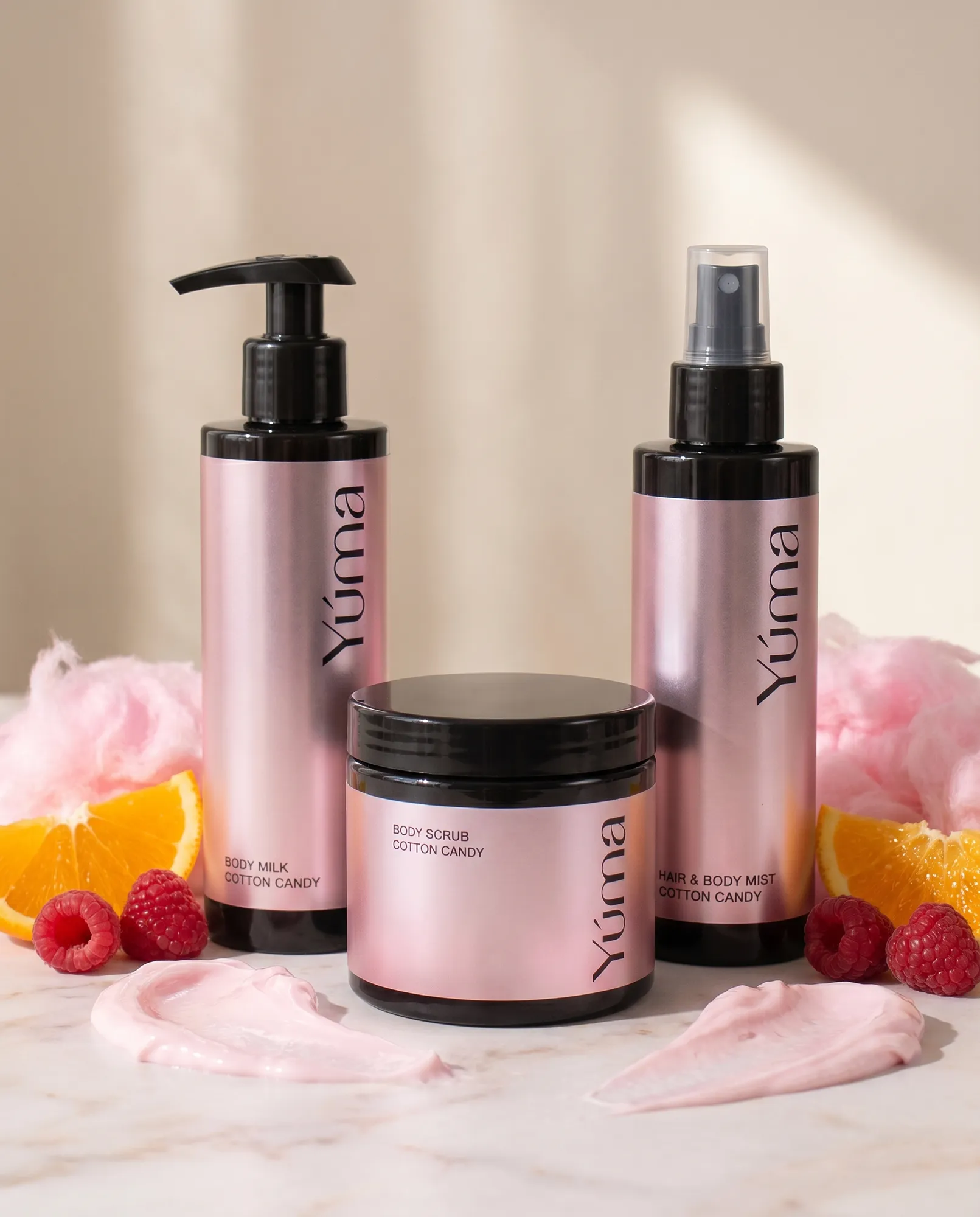 Three pink metallic YUMA Cotton Candy bottles surrounded by raspberries, oranges, and pink cotton candy.
