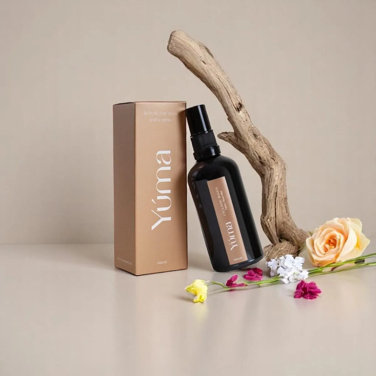 YUMA Cycladic Roots Fabric Mist in a dark bottle, next to its paper packaging on wood with flowers.