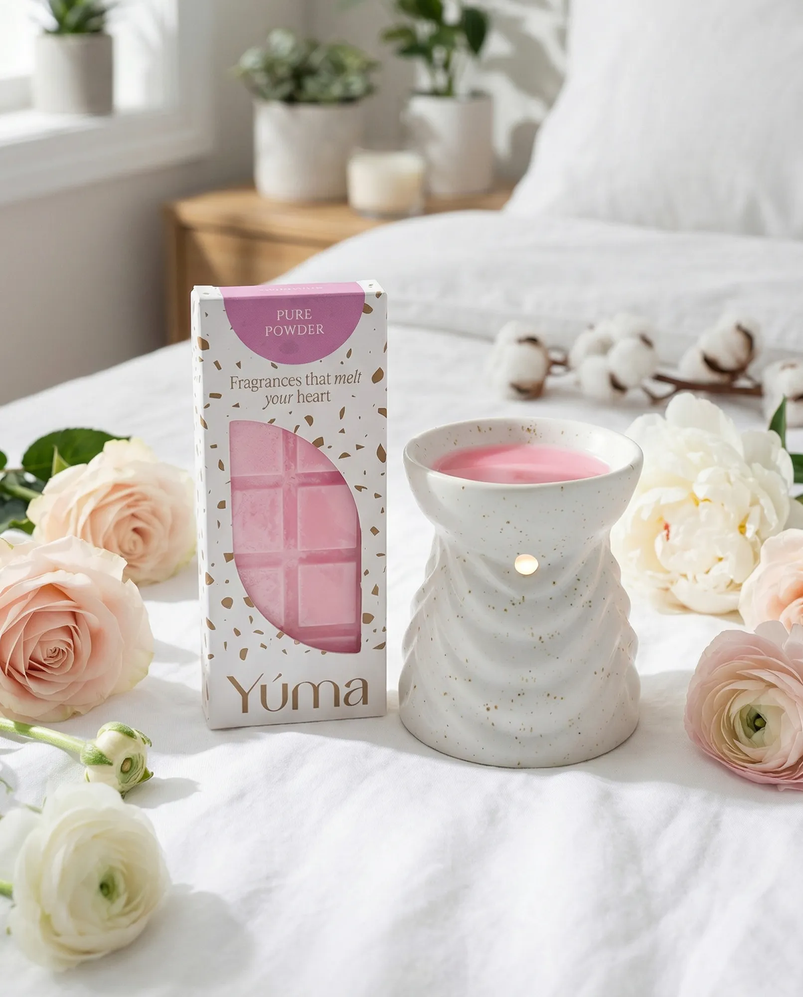 YUMA Pure Powder pink wax melts beside a ceramic wax warmer and roses on a white bed.