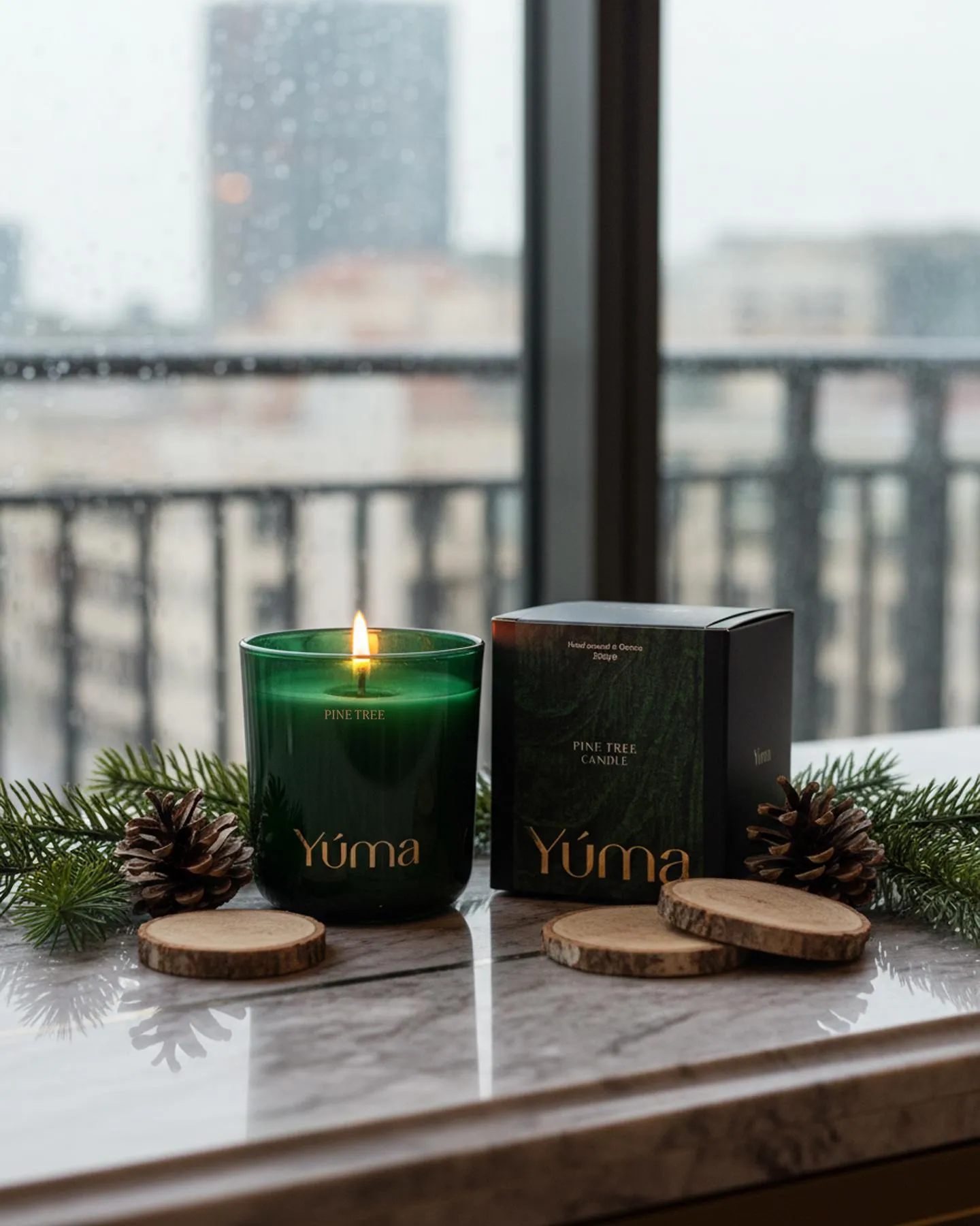 Lit YUMA Pine Tree candle in a green glass vessel beside its packaging on marble.