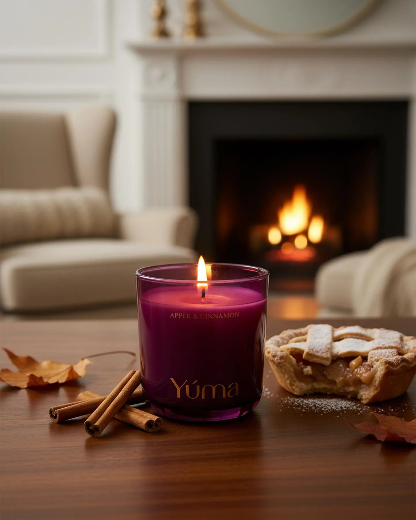 Lit YUMA Apple Cinnamon candle beside apple pie and cinnamon sticks in front of a fireplace.