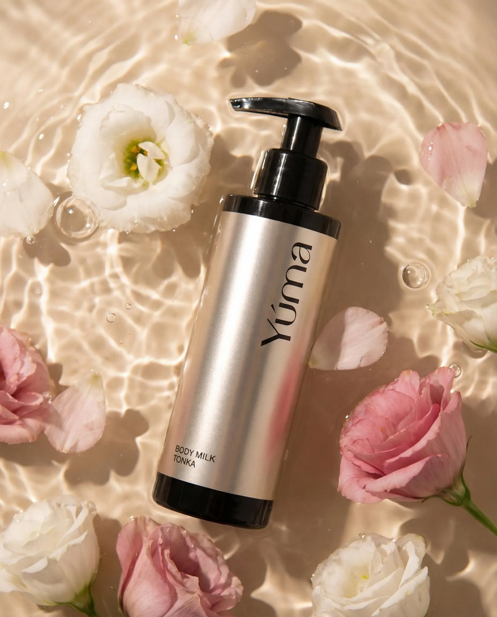 YUMA Tonka Body Milk in a metallic bottle submerged in water with pink and white flowers.
