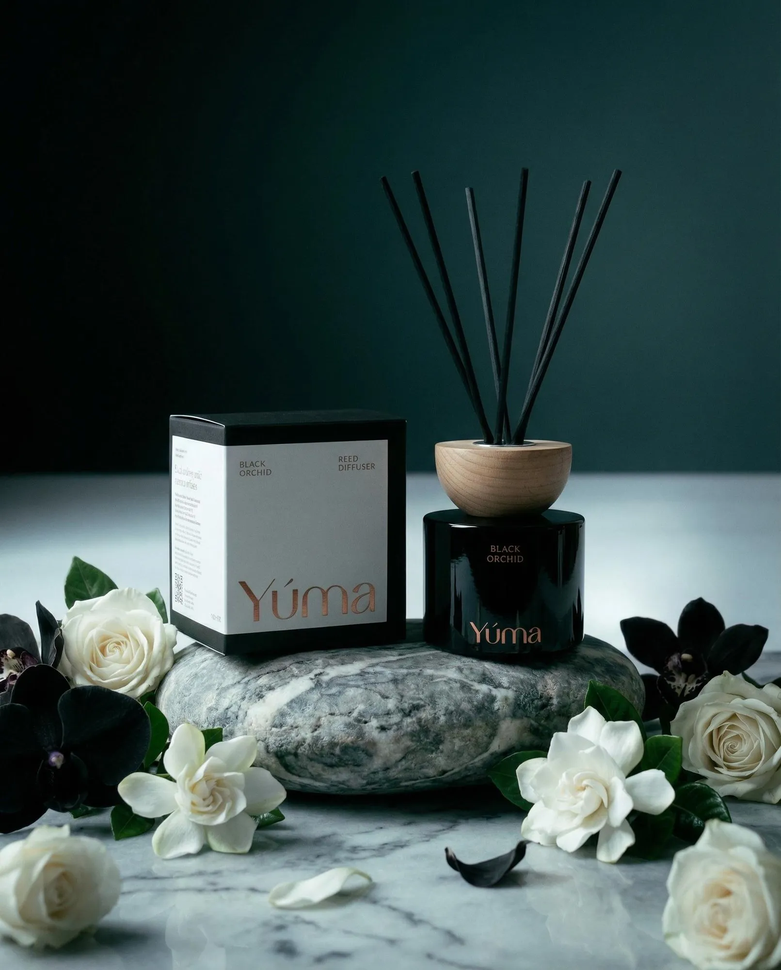YUMA Black Orchid reed diffuser next to its packaging, placed on a stone surface with white flowers.