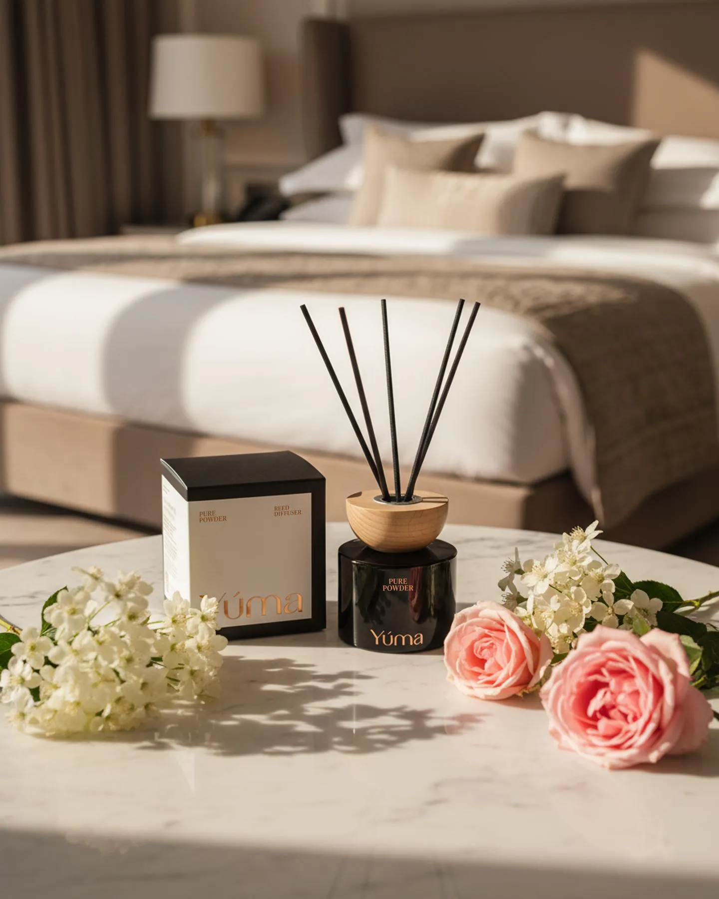 YUMA Pure Powder reed diffuser with black reeds beside pink roses on a marble table.