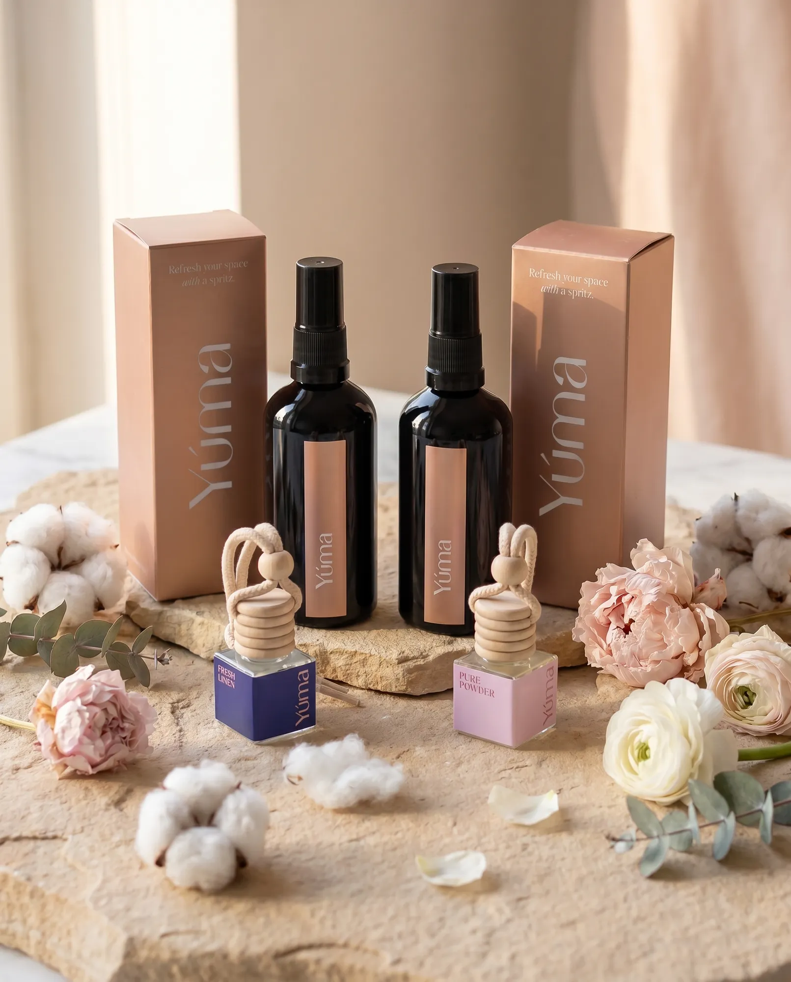 YUMA Fresh Linen and Pure Powder Fabric/Room Mists with their two matching Car Diffusers among flowers.