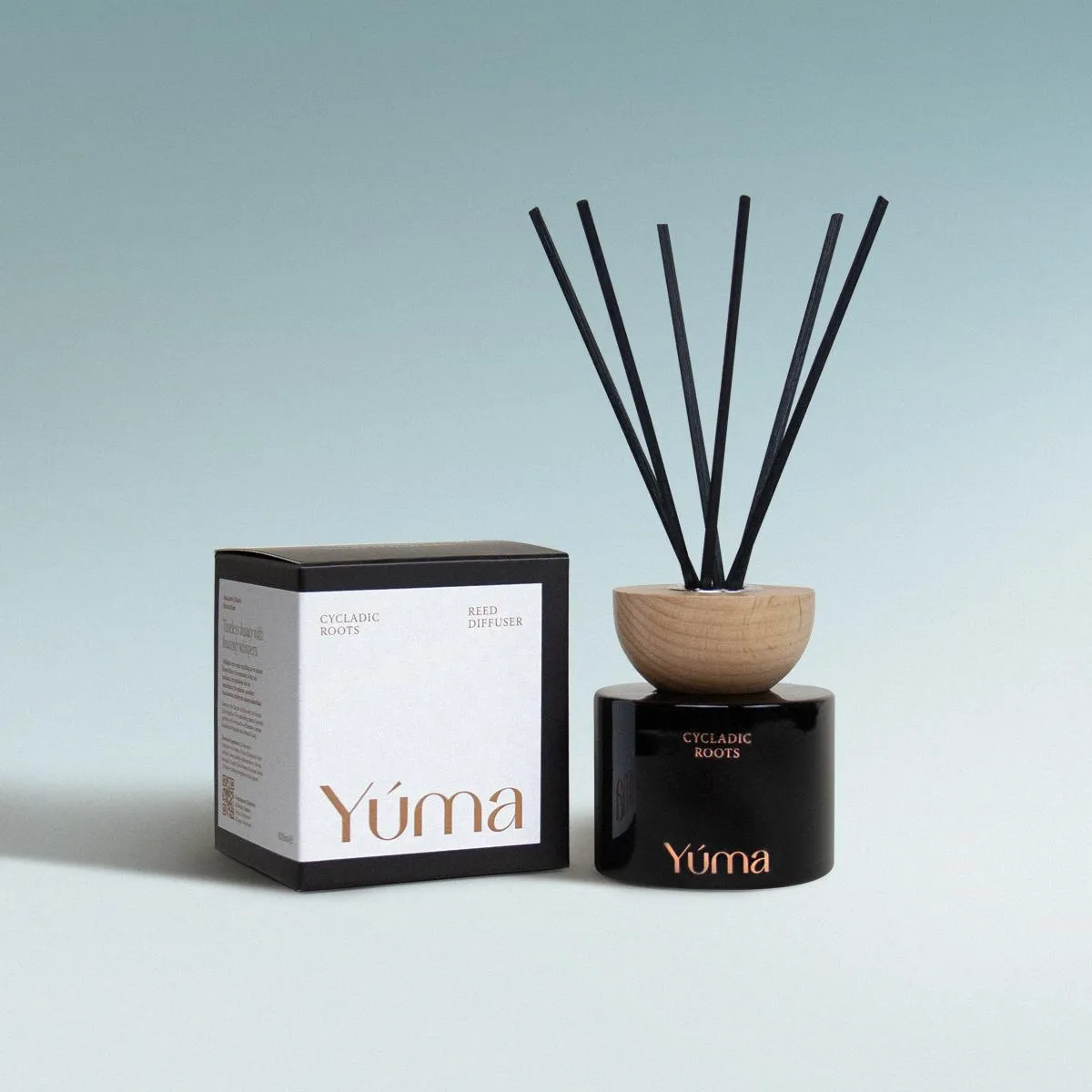 YUMA Cycladic Roots reed diffuser in a black glass bottle with a wooden cap, next to its packaging.