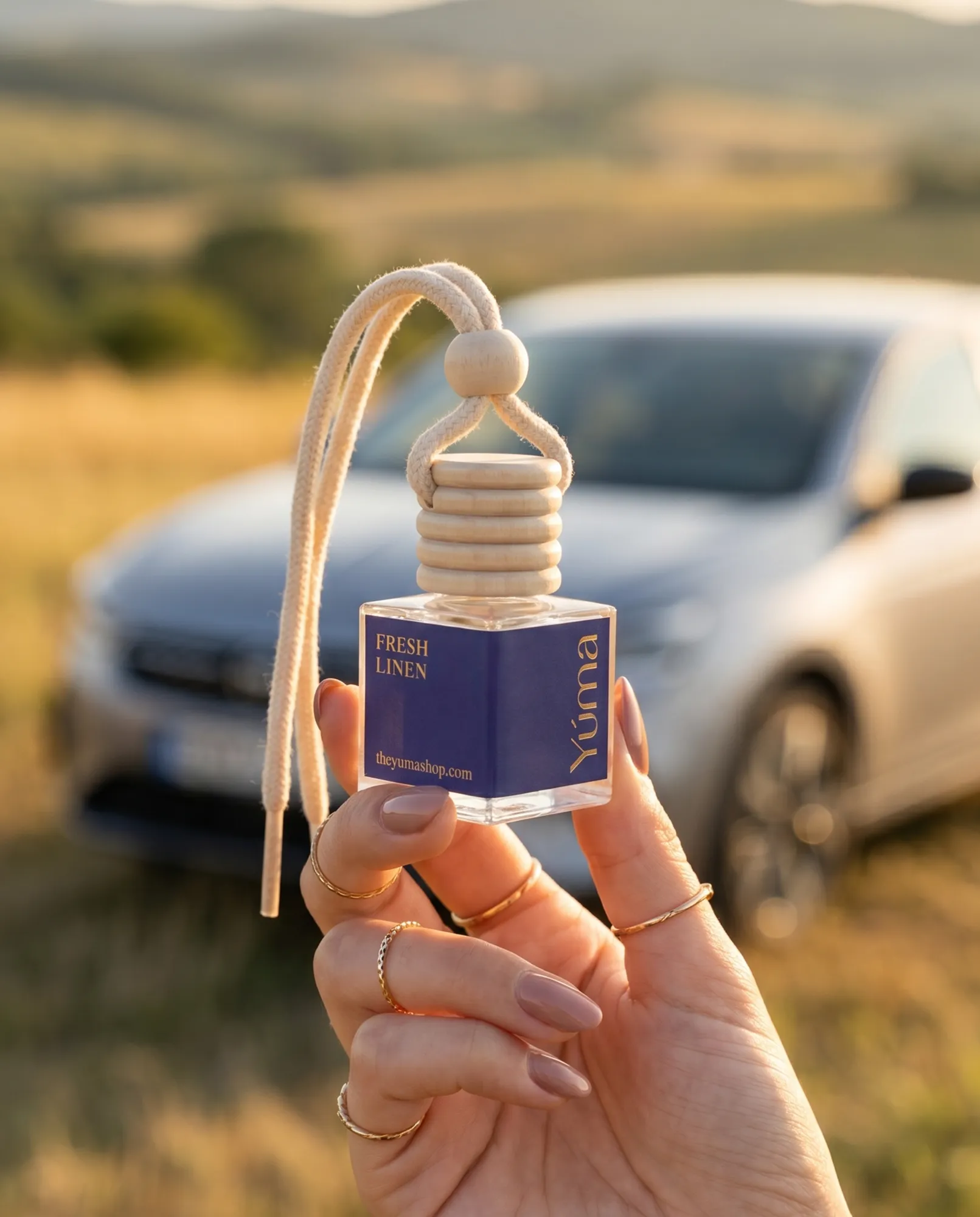 A woman's hand holding the YUMA Fresh Linen Car Diffuser, with a car in a natural outdoor setting in the background.