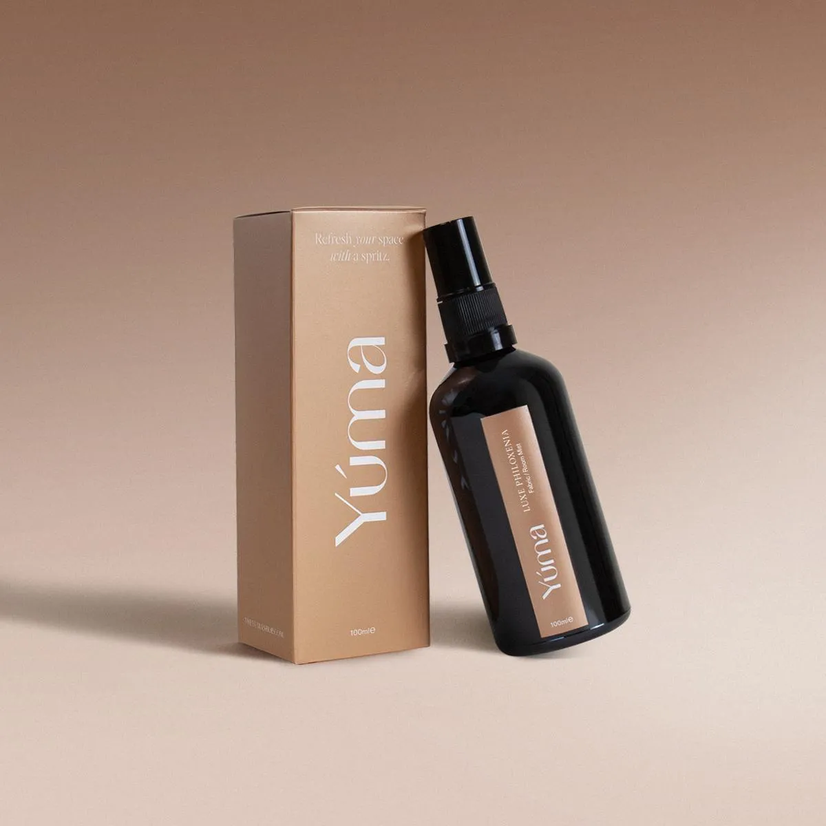 YUMA Luxe Philoxenia dark glass spray bottle beside its beige paper packaging on a neutral background.