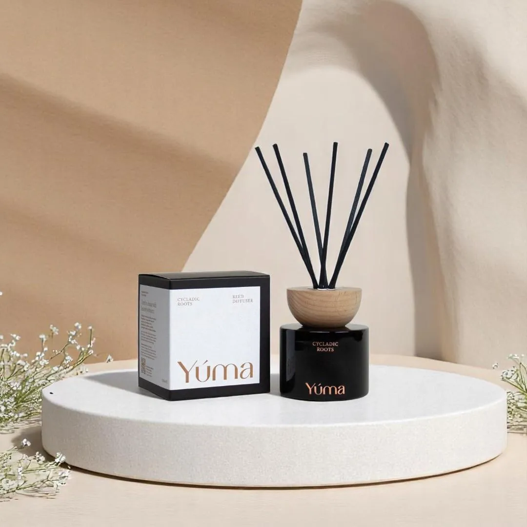 YUMA Cycladic Roots reed diffuser with black sticks, placed next to its packaging on a pedestal.
