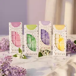 Signature Collection - Wax Melt Set Four YUMA wax melts in paper packaging on marble, surrounded by lilac and jasmine blossoms.