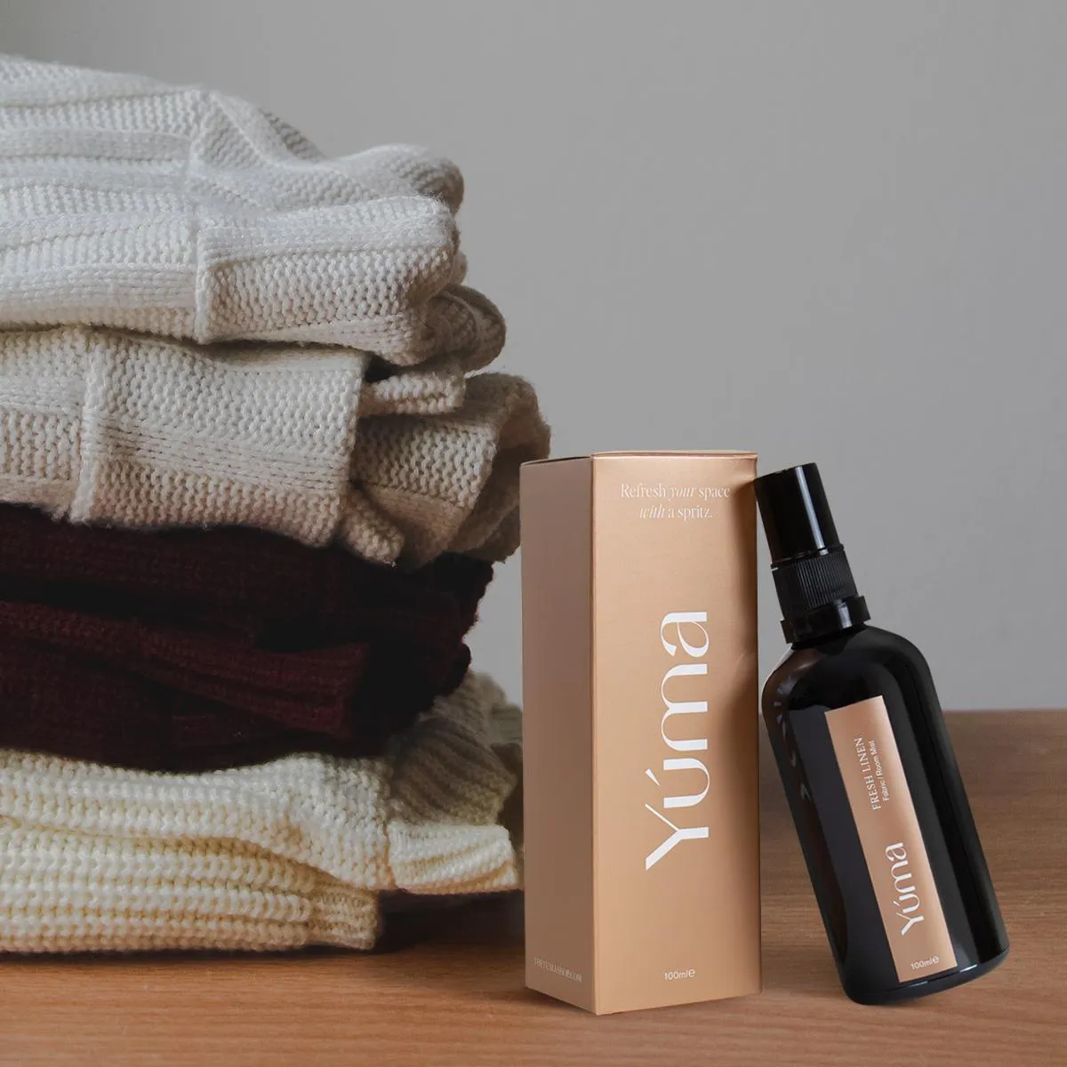 Fabric Mist - Velvet Cashmere - Scented Fabric Spray Dark YUMA Fabric Mist bottle next to a stack of knit fabrics on a wooden surface.
